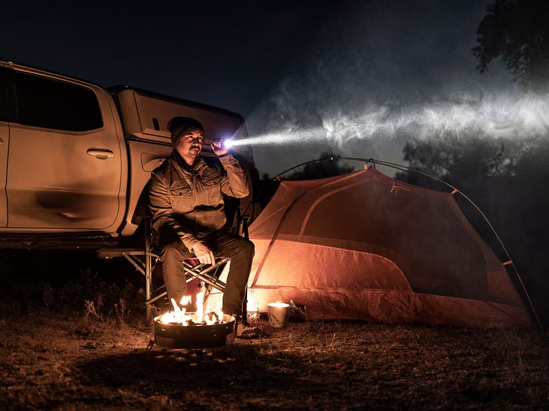 Man sitting beside a campfire at a nighttime campsite, using a flashlight to illuminate the area near his tent and pickup truck.